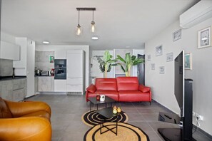 Ein modernes Wohnzimmer mit einem roten Sofa, einem Couchtisch und einem Fernseher. Im Hintergrund ist eine Küche mit weißen Schränken und einer Arbeitsplatte zu sehen.