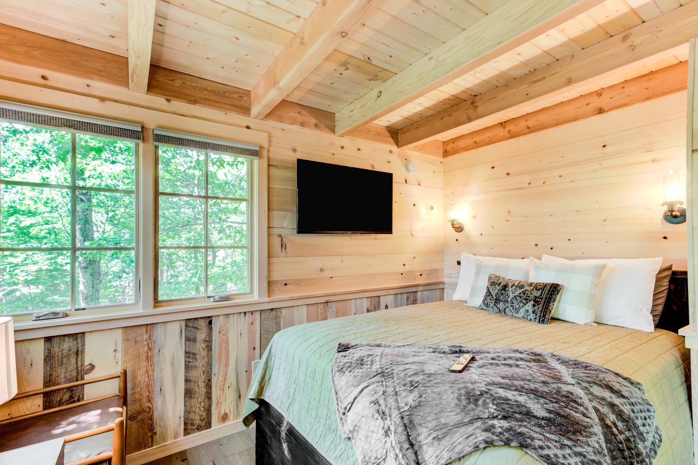 Een slaapkamer in een houten chalet met een bed, een wandtelevisie en een raam met uitzicht op bomen.