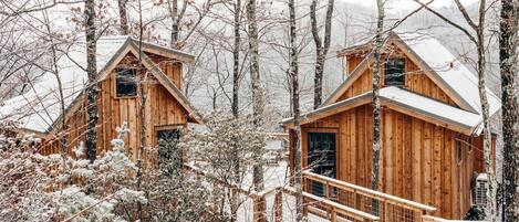 Deux cabanes en bois, dont les toits sont recouverts de neige, entourées d’arbres et avec une chaîne de montagnes en arrière-plan.