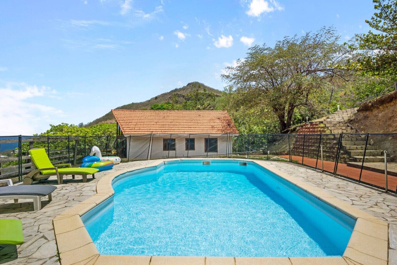 A swimming pool with a tiled deck, surrounded by lounge chairs and a small building with a tiled roof.