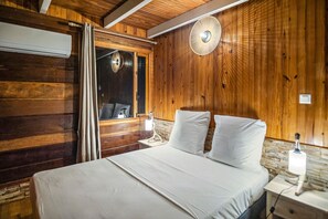 A wooden cabin-style room with a bed, a window with curtains, a wall-mounted fan, and a nightstand with a lamp. - Chalet Jujube - Beautiful cottage with a pool (Les Anses-d`Arlet)