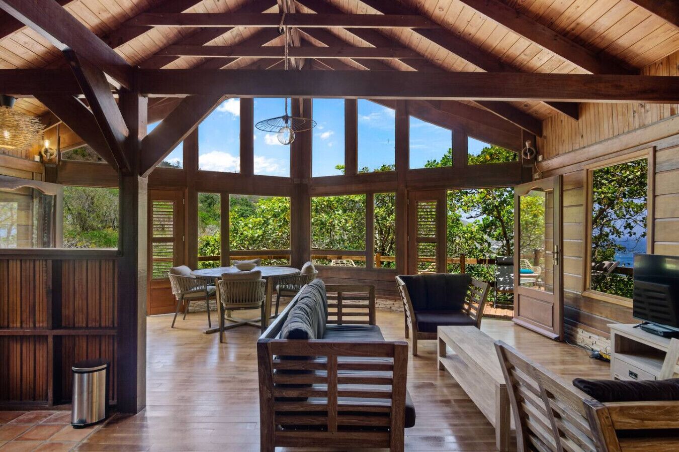 A covered outdoor seating area with wooden furniture, large windows, and a view of greenery.