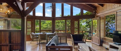 A covered outdoor seating area with wooden furniture, large windows, and a view of greenery.