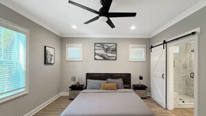 Quarto com cama, ventoinha no teto, mesa de cabeceira, quadro emoldurado, janela e porta deslizante para área de chuveiro.