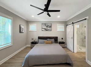 Un dormitorio con una cama, ventilador de techo, mesita de noche, cuadro enmarcado y ventana. Puerta corrediza que da a una zona de ducha.