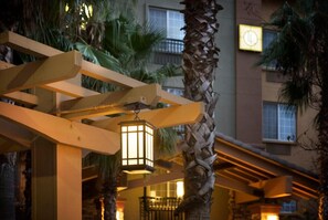 Eine hölzerne Pergola mit hängenden Laternen, Palmen und einer Gebäudefassade im Hintergrund.