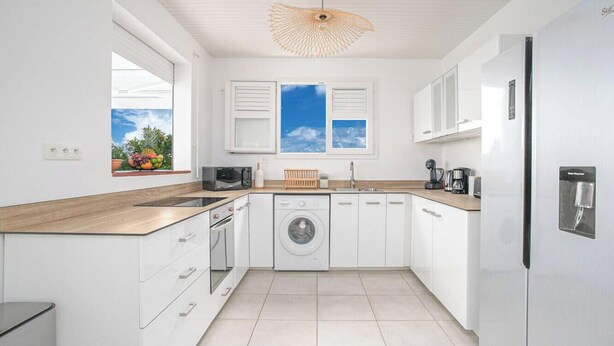 A modern kitchen with white cabinetry, a built-in washing machine, and a large window with a view of the sky. - Villa Kaliemma - Tropical Villa in Martinique (Le Marin)