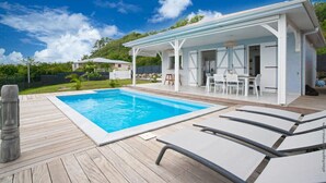 A swimming pool with a wooden deck, a covered outdoor seating area, and a building with white walls and a flat roof. - Villa Kaliemma - Tropical Villa in Martinique (Le Marin)