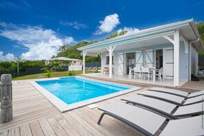 A swimming pool with a wooden deck, a covered outdoor seating area, and a building with white walls and a flat roof.