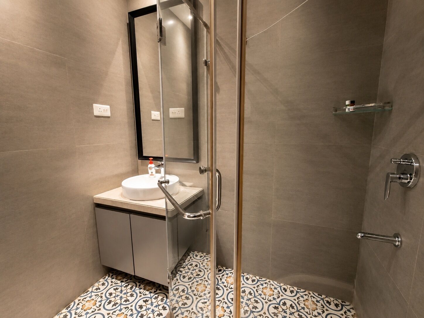 A modern bathroom with a glass shower enclosure, a white sink with a mirror, and patterned floor tiles.