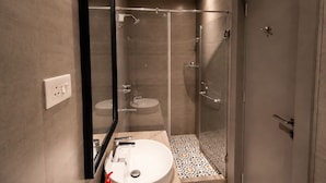 A modern bathroom with a glass-enclosed shower, a sink with a mirror, and a tiled floor. - The Bay Suites - Salt Lake AE Market (Bidhannagar)
