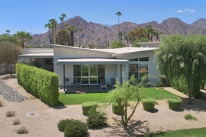 A modern house with a patio, surrounded by a well-maintained garden and palm trees, with mountains in the background. - 4BR| Pool | Firepit| Patios, Mtn Views| Next to Tennis Garden & Golf Resort (Indian Wells)