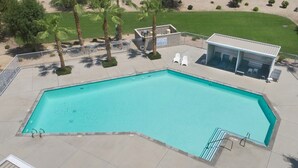 Uma piscina retangular com um deck revestido de azulejos, cercada por uma cerca de metal, palmeiras e um pequeno edifício com telhado.