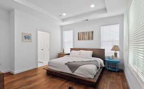 A bedroom with a wooden bed, white walls, a wooden floor, a nightstand with a lamp, and a door. - Millville by the Sea- shuttle access Bethany beach (Millville)