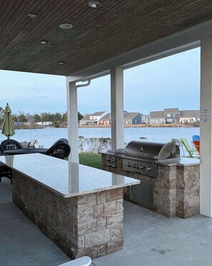 An outdoor kitchen with a stone countertop and grill, overlooking a body of water and residential buildings. - Millville by the Sea- shuttle access Bethany beach (Millville)