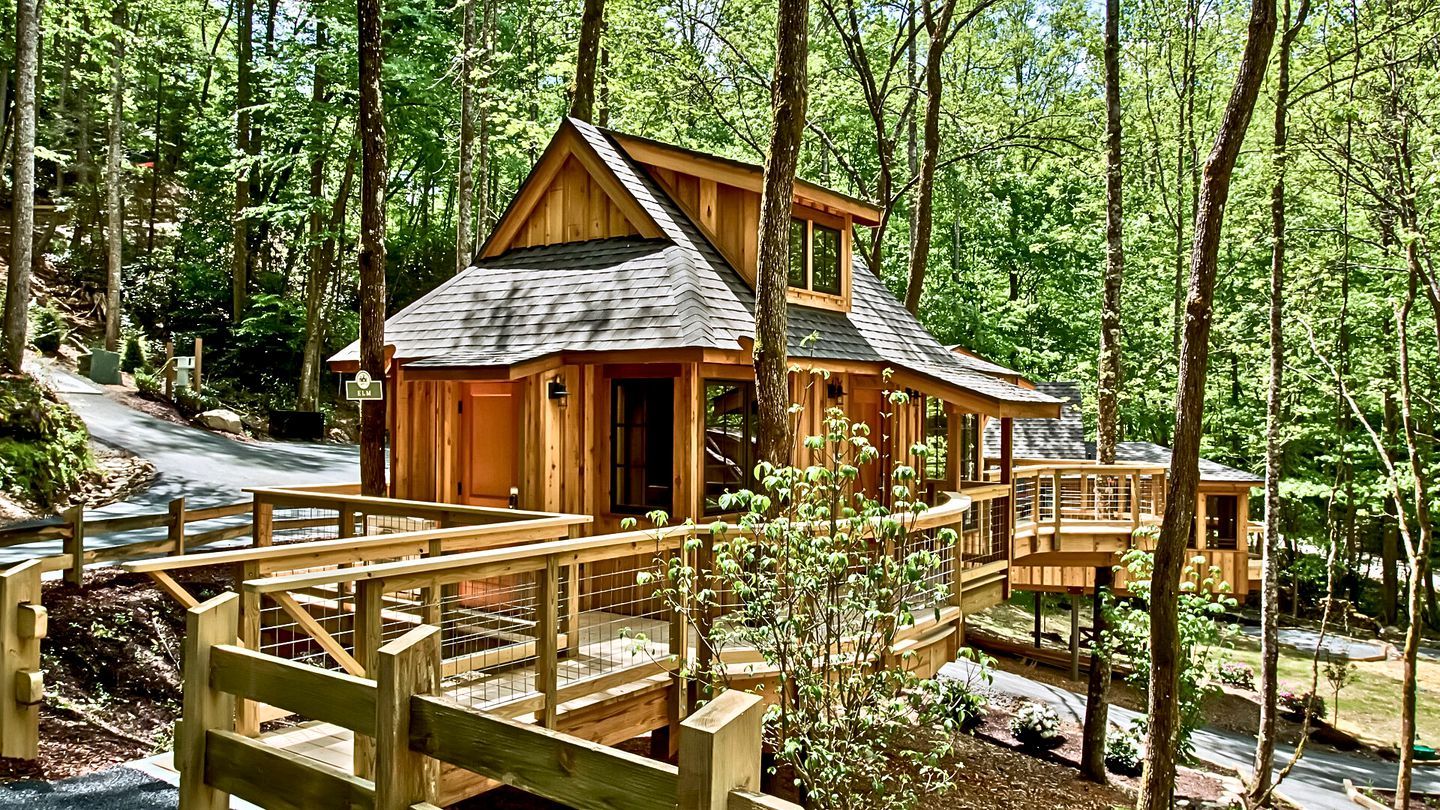 A wooden cabin with a deck and a small bridge, surrounded by a forest.