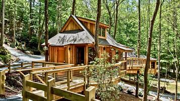 A wooden cabin with a deck and a small bridge, surrounded by a forest.