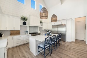A spacious kitchen with white cabinetry, a central island with bar stools, a stainless steel refrigerator, and hanging wicker pendant lights. - Seas & Sunsets 201 LAK (Port Aransas)