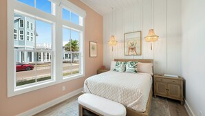 A bedroom with a large window, a bed with a quilt, two bedside tables, and a bench. - Seas & Sunsets 201 LAK (Port Aransas)