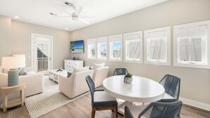 A modern living room with a white sofa, a round dining table, wicker chairs, a ceiling fan, and a view of the beach through large windows. - Seas & Sunsets 201 LAK (Port Aransas)