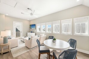 Ein modernes Wohnzimmer mit einem weißen Sofa, einem runden Esstisch, Korbstühlen und einem Deckenventilator, mit Blick auf den Strand durch große Fenster.