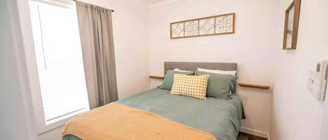 A bedroom with a bed, a window with curtains, a wall-mounted artwork, and a bedside table.