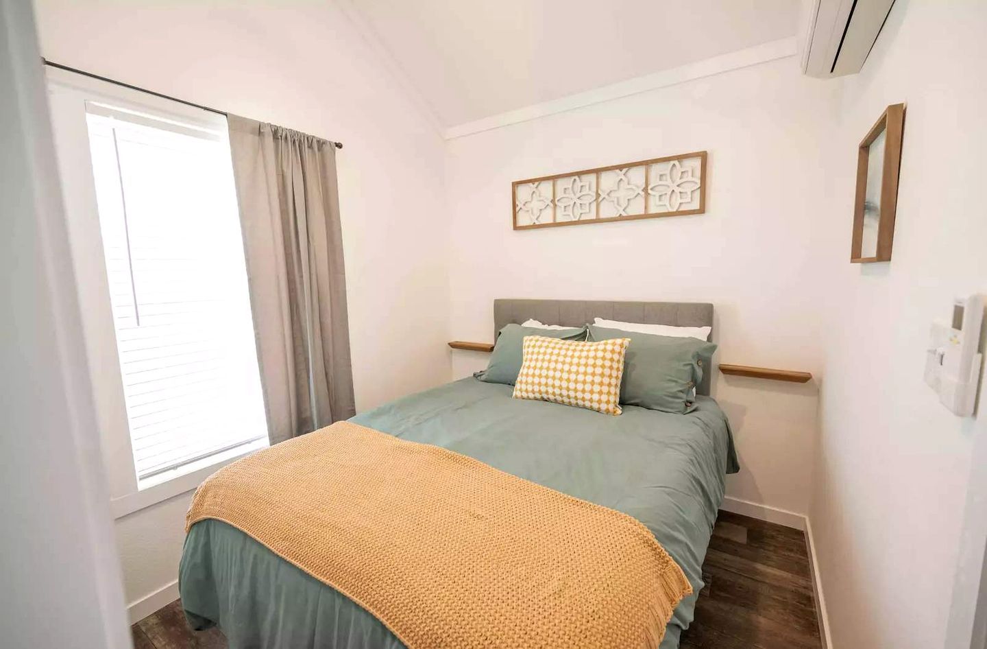 A bedroom with a bed, a window with curtains, a wall-mounted artwork, and a bedside table.