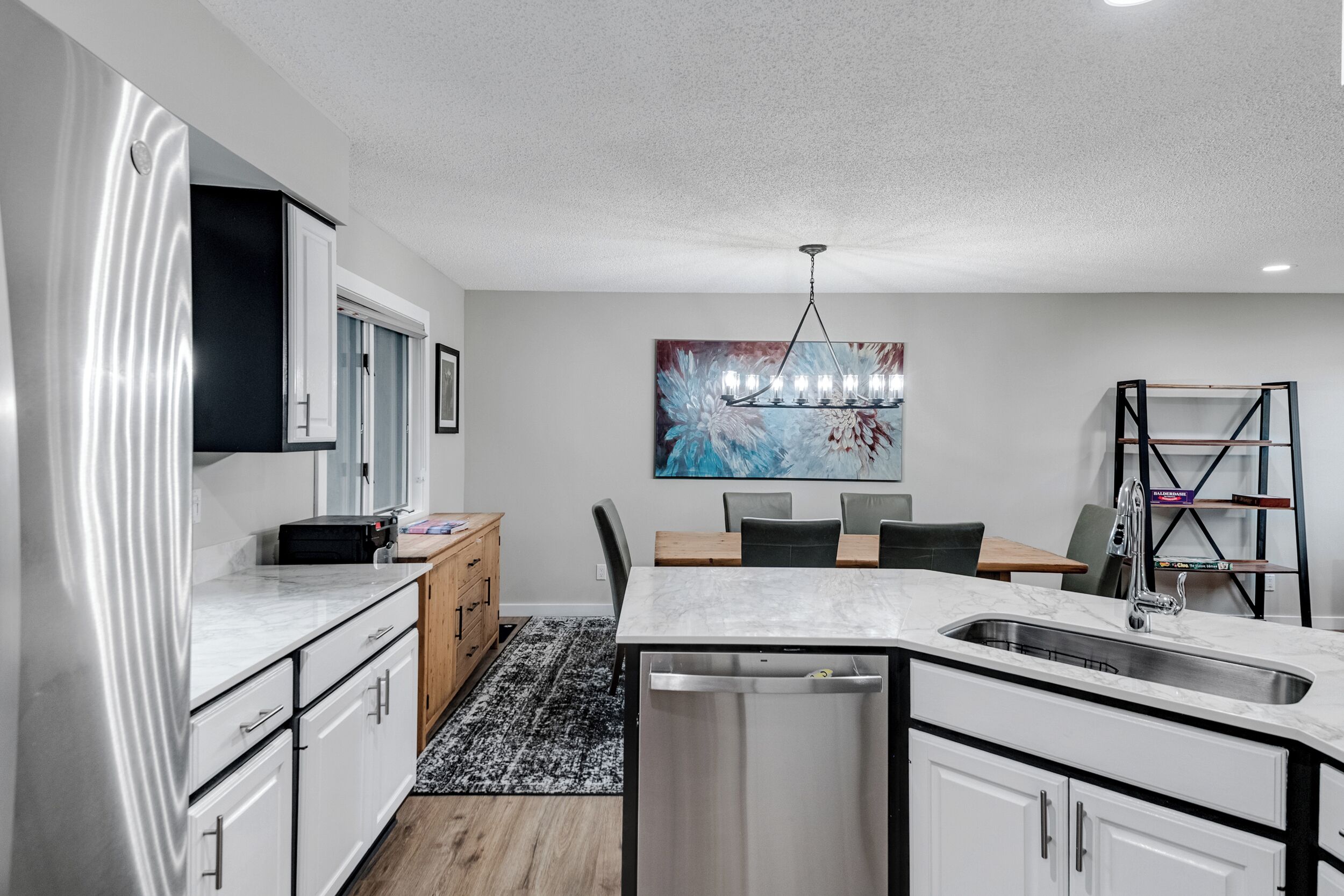 A modern kitchen with white cabinetry, a marble countertop, a built-in dishwasher, and a dining area with a table and chairs.