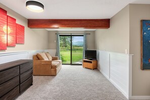A living room with a sofa, TV, and a view of a green landscape through a glass door.