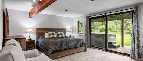 A bedroom with a large bed, a sofa, a nightstand, a lamp, and a view of a garden through a sliding glass door.