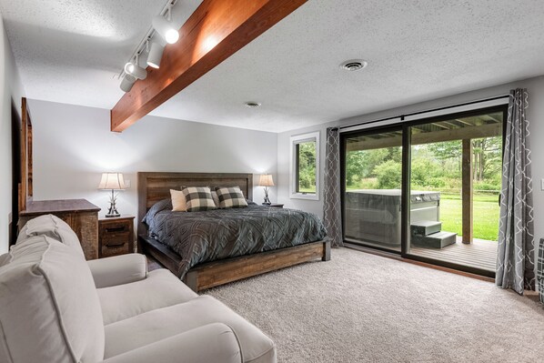 A bedroom with a large bed, a sofa, a nightstand, a lamp, and a view of a garden through a sliding glass door.