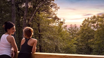 Two individuals on a wooden deck overlooking a forested area with a sunset in the background.