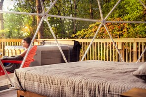 Una silla hamaca con una persona sentada, una cama con una manta y una terraza de madera con una valla.