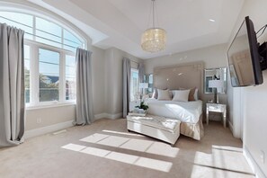 Ein modernes Schlafzimmer mit einem großen Bett, einem Nachttisch, einem Beistelltisch, einem an der Wand montierten Fernseher und einer Kristallleuchtre.