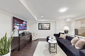Ein modernes Wohnzimmer mit Flachbildfernseher, dunklem Holzboden, einer dunklen Couch, einem weißen Teppich und einer Zimmerpflanze.
