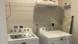Ein Waschbereich mit zwei weißen Maytag Waschmaschinen und Trocknern, einem Metallregal mit Wäschezubehör und einem Bügeleisen, das hängt.