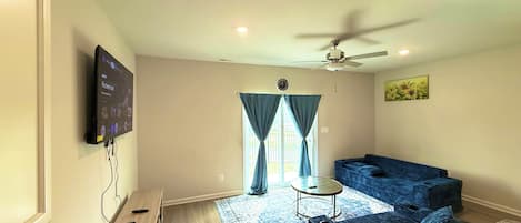 Una sala de estar con un televisor de pantalla plana montado en la pared, un ventilador de techo, un sofá, una mesita redonda y una ventana con cortinas.