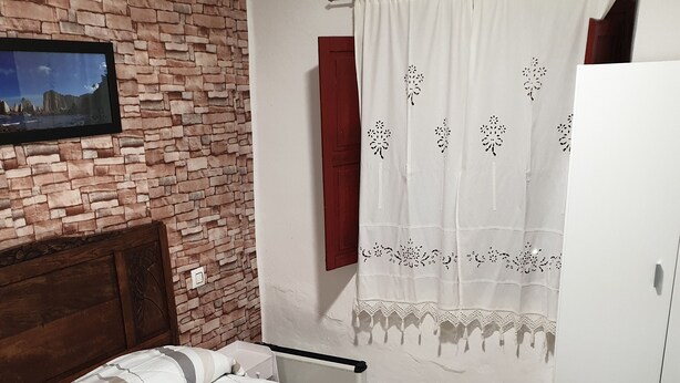 Ein Schlafzimmer mit Backsteinwand, einem Bett mit gemusterten Betttextilien, einem Holz-Kopfteil, einem weißen Kleiderschrank und einem Fenster mit weißem Vorhang.