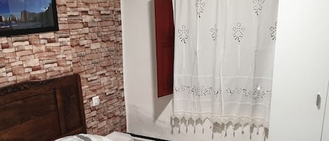 A bedroom with a brick wall, a bed with patterned bedding, a wooden headboard, a white wardrobe, and a window with a white curtain.