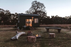 A small cabin with a glass door entrance, a fire pit, and a bench in a grassy area.