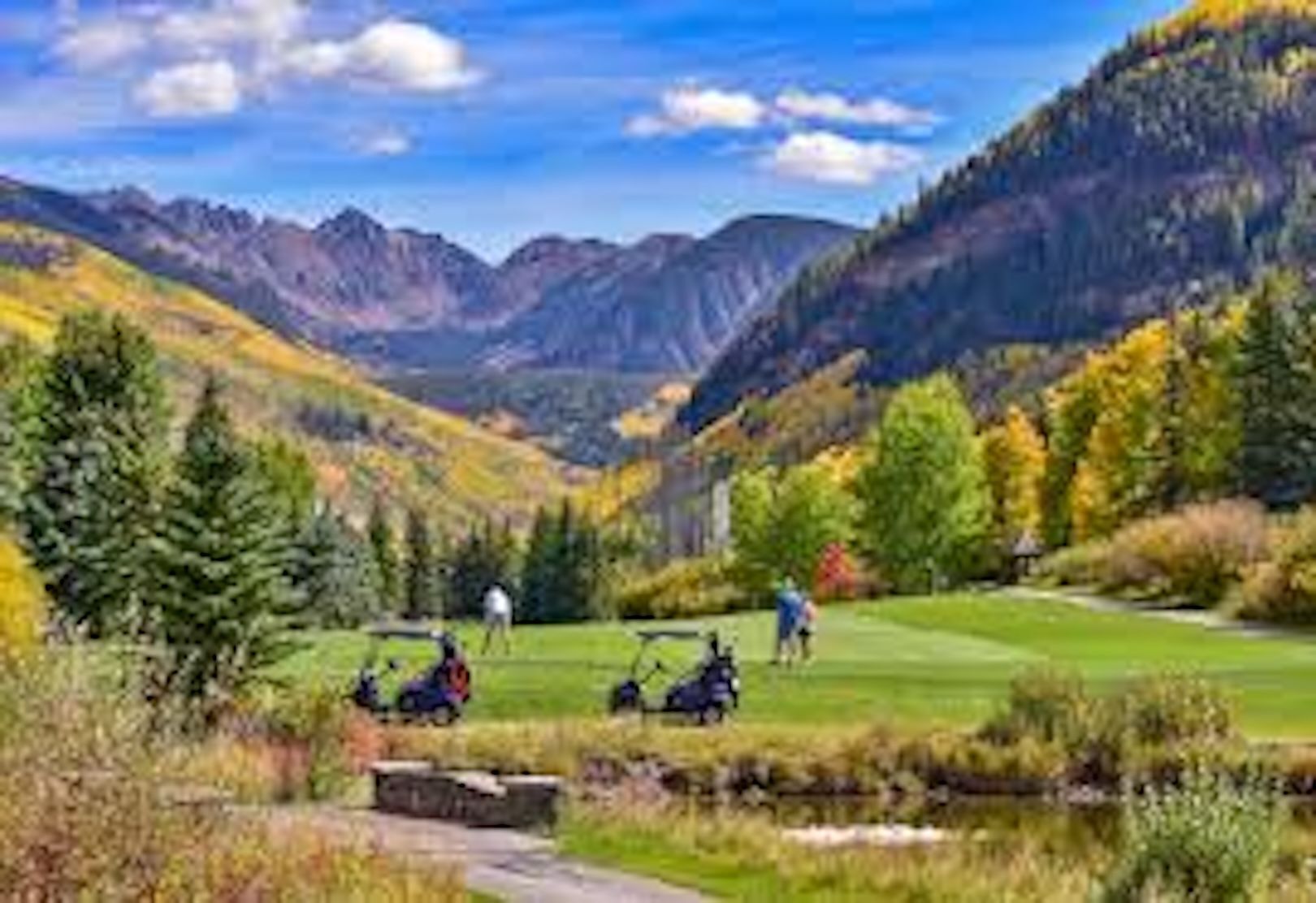 Golfers on a mountain course surrounded by autumn trees and high peaks.
