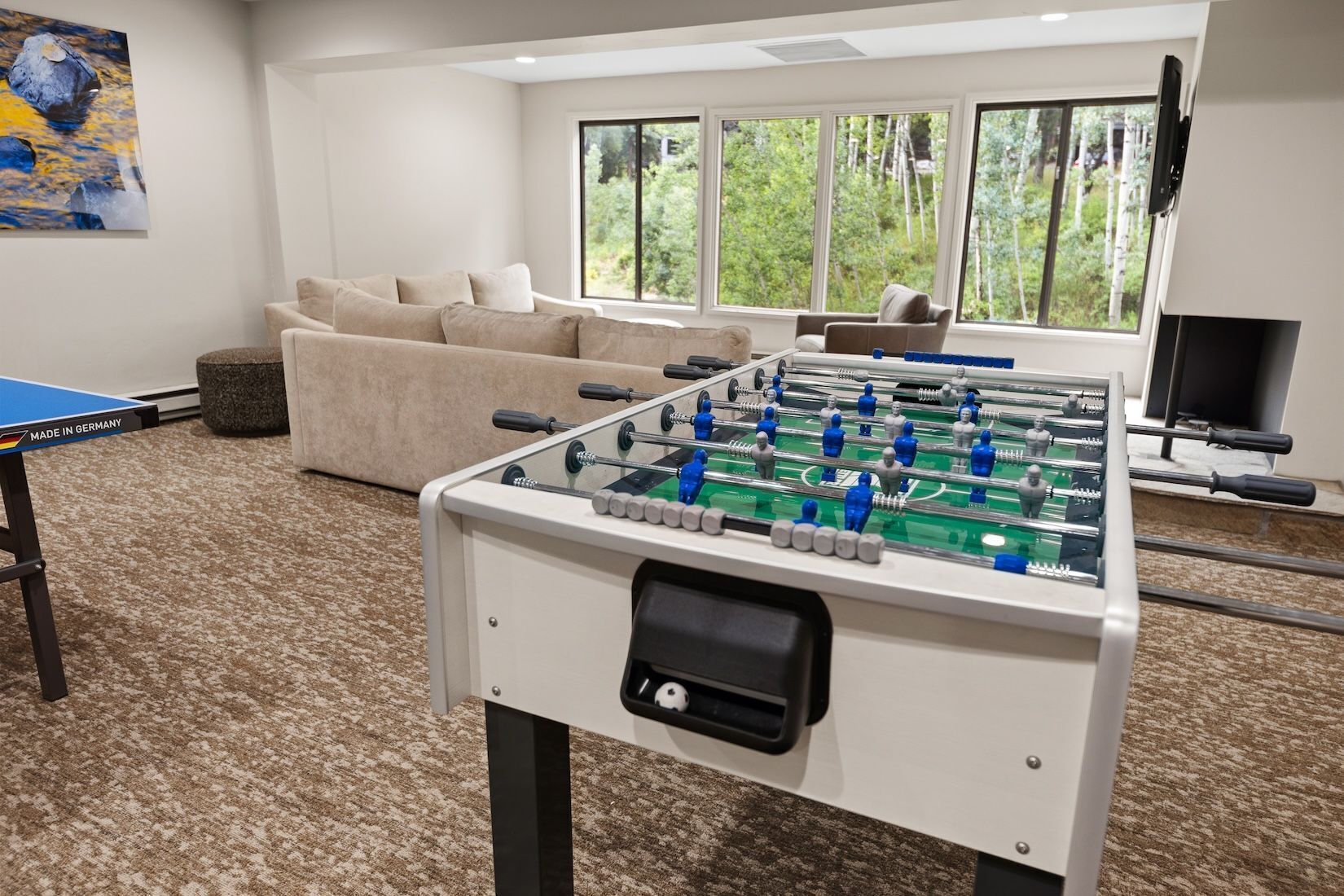 Habitació amb taula de futbolí, sofà, televisió i finestra amb vista a arbres.