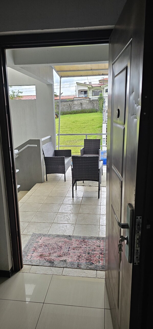 Vista a través de una puerta abierta que da a un espacio exterior cubierto con suelo de baldosas, dos sillas de mimbre y un tapete estampado.