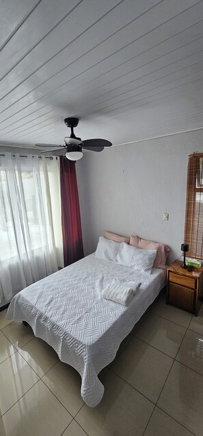 Un dormitorio con cama, ventilador de techo, mesita de noche, lámpara y ventana con cortinas.