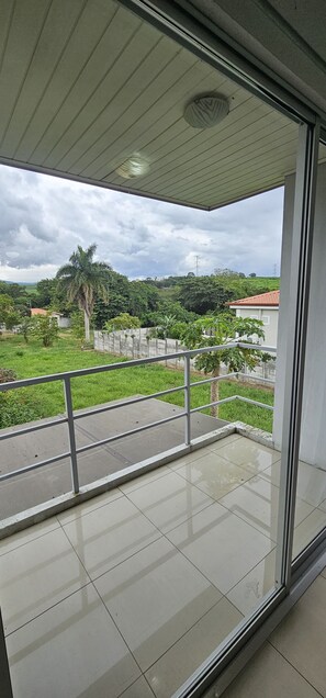 Un balcón con barandilla de metal, piso de baldosas y vista a un paisaje verde con árboles y una colina.