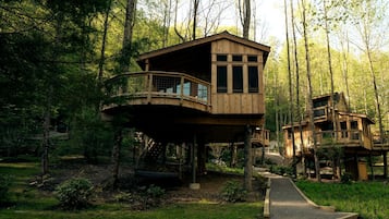 A wooden treehouse structure with a balcony, surrounded by a forested area.