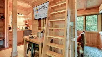 A cozy wooden cabin interior with a kitchen area, a dining table with a bottle and glasses, and a sign for 'AYRES Vacation Rentals.'