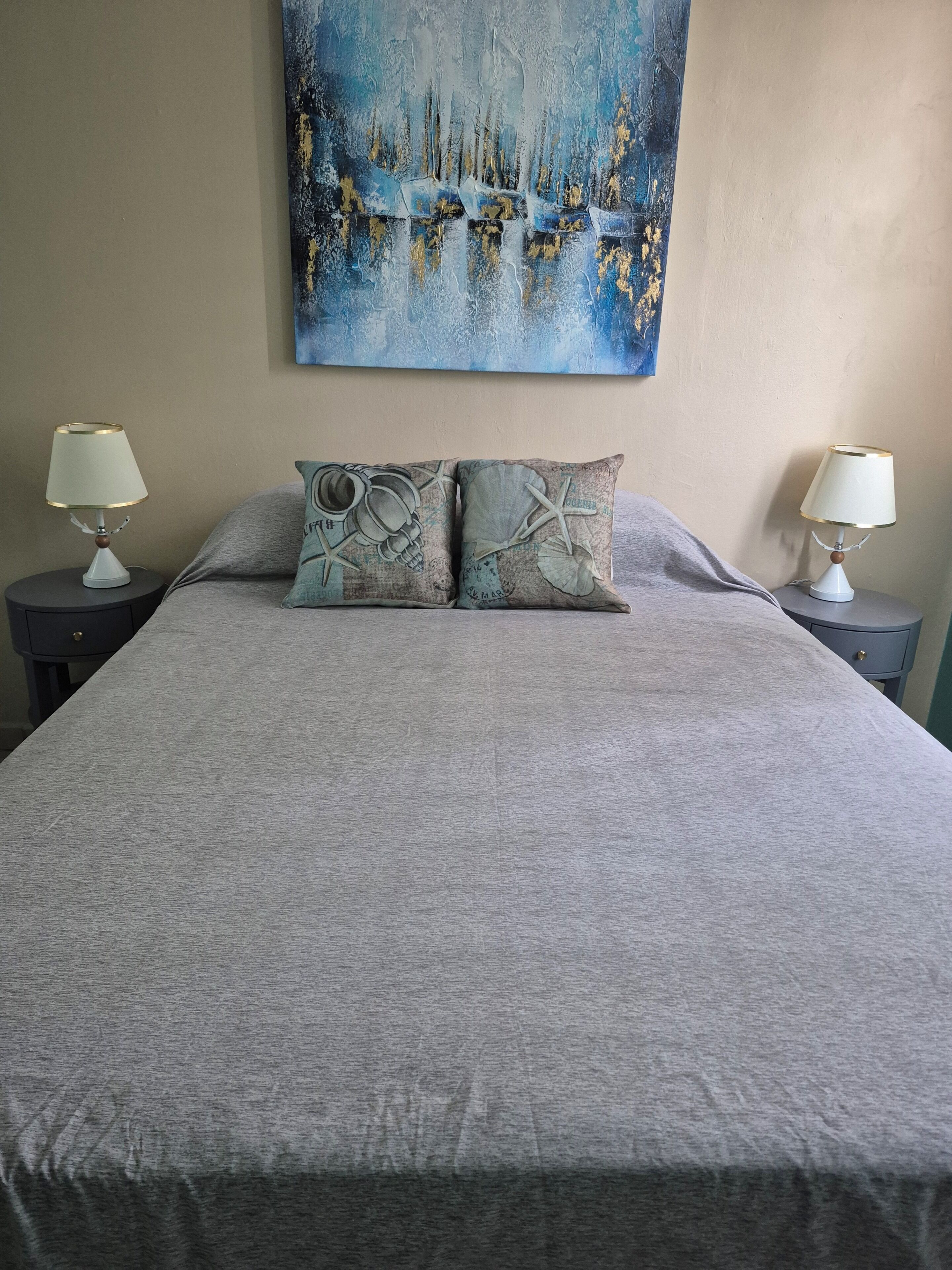 A bed with a grey bedspread, two pillows with nautical designs, and two bedside lamps on matching nightstands.
