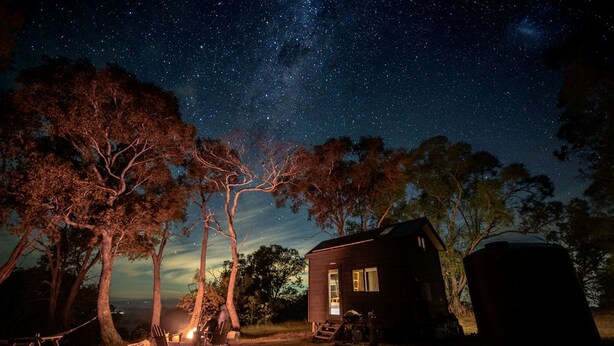 Una cabaña pequeña bajo un cielo estrellado, rodeada de árboles y equipo de campamento.