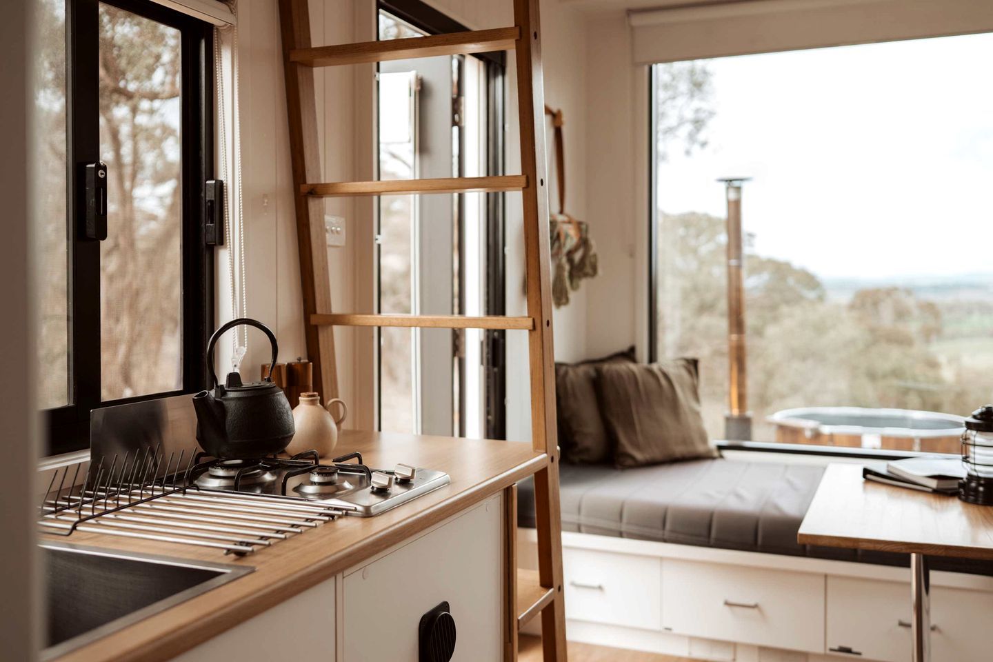 A kitchen with a stove, kettle, and utensils, a wooden ladder, and a view of the outdoors through large windows.